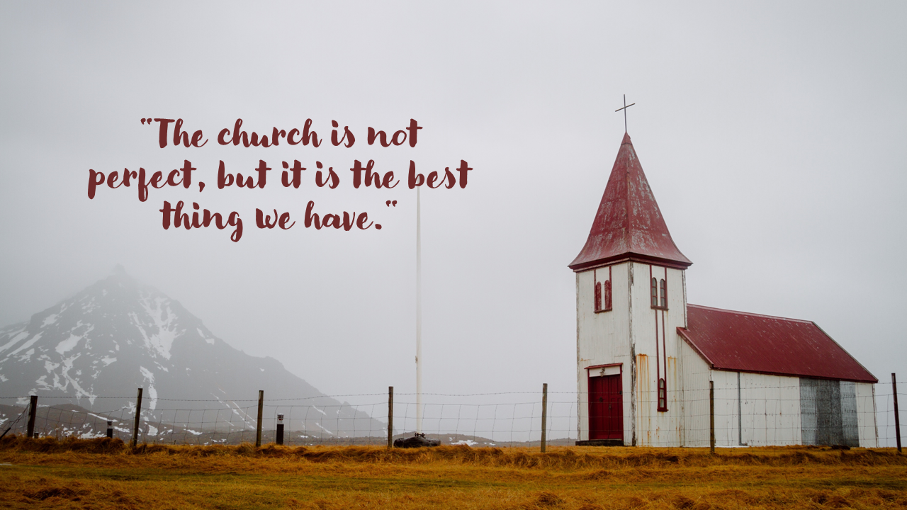 A white church with a red roof, standing alone against a foggy sky and snow capped mountains, surrounded by a fence.