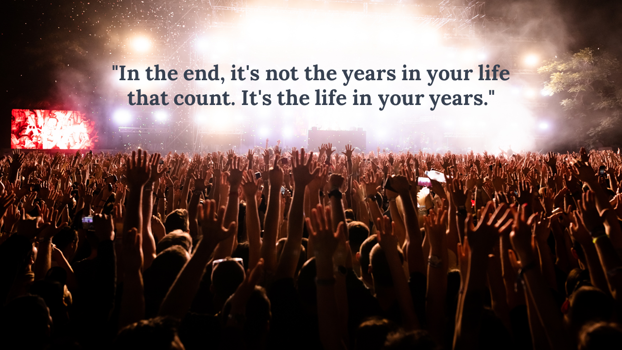 A large crowd with many hands raised is silhouetted against bright stage lights and haze at a concert or event.